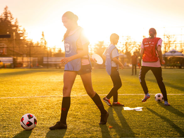 Young girls in pinnies playing soccer on a field as a golden sun shines.