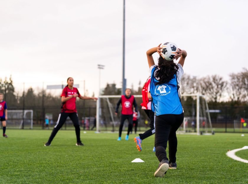 Young girls in pinnies playing soccer on a field as the ball is being thrown in – clicking this link will open a larger version of it in the browser window.