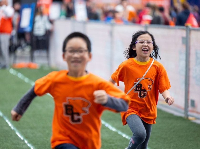 Two young children in BC Lions shirts race down a field during a kids event at the 2024 Grey Cup Festival – clicking this link will open a larger version of it in the browser window.