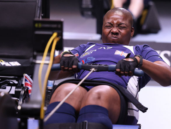 A female athlete representing the United Kingdom strenuously participates in an indoor rowing event.